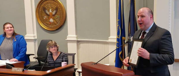 Two women with the county seal on the wall behind them look on while a man at a podium holds up a proclamation he has just received